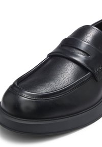 Μαύρο δερμάτινο μοκασίνι με διάτρητο σχέδιο στο επάνω μέρος, στρογγυλεμένη μύτη και παχύ λαστιχένιο πέλμα, εμφανιζόμενο σε κοντινή λήψη.