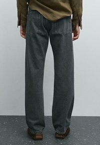 Jeans di denim grigio scuro con una vestibilità ampia, dotati di tasche posteriori e una texture leggermente logora. Abbinati a scarpe marroni.
