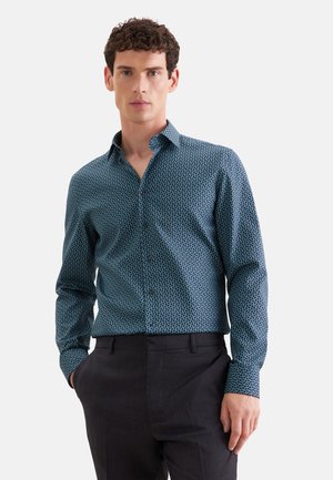 Mann trägt ein blau gemustertes, langärmeliges Hemd mit schwarzen Hosen, steht mit einer Hand in der Tasche vor einem einfarbigen Hintergrund.