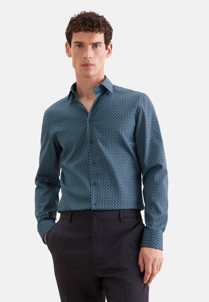 Homme portant une chemise bleue à motifs à manches longues avec un pantalon noir, debout avec une main dans la poche devant un fond uni.