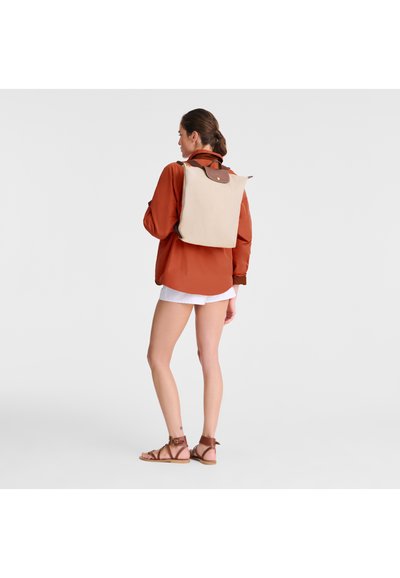 Brauner Rucksack mit Lederakzenten, rechteckige Form, verstellbare Riemen, getragen über einem orangefarbenen Shirt und weißen Shorts.