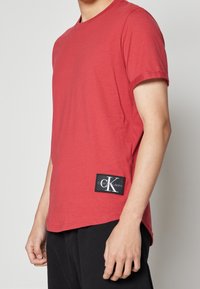Camiseta de algodón roja con mangas cortas, escote redondo y un parche negro con el logo de Calvin Klein Jeans en la parte inferior izquierda.