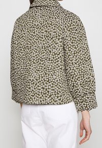 Padded jakke med olivengrøn baggrund og et hvidt blomsterprint. Designet har en høj krave og rynkede ærmer.