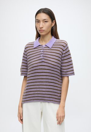 Frau trägt ein kurzärmeliges, lila-braun gestreiftes Poloshirt mit Knöpfen, kombiniert mit weißen Hosen vor einem schlichten weißen Hintergrund.