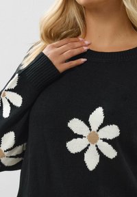 Schwarzer Strickpullover mit großen weißen Gänseblümchenmustern und beigen Mittelkreisen an den Ärmeln. Rundhalsausschnitt und gerippte Bündchen.