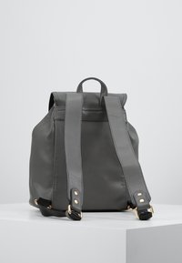 Sac à dos en cuir gris de forme arrondie, avec deux bretelles, une poignée supérieure et des accessoires en métal doré. Texture lisse avec un minimum de détails.