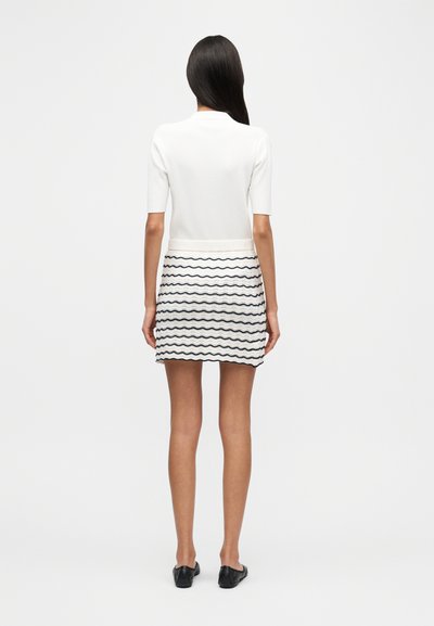 Σετ δύο τεμαχίων που αποτελείται από λευκό ριμπ πλεκτό τοπ με κοντά μανίκια και φούστα με κυματιστό ασπρόμαυρο σχέδιο. Μαύρα παπούτσια.