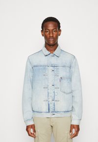 Chaqueta de mezclilla azul claro con cierre de botones en la parte delantera, dos bolsillos en el pecho y un cuello clásico. Acentos desgastados en toda la tela.