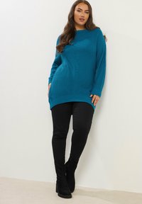 Turkusowy sweter prążkowany z długim rękawem, bocznymi kieszeniami i lekko zaokrąglonym dołem, zestawiony z czarnymi rurkami i czarnymi botkami za kostkę.