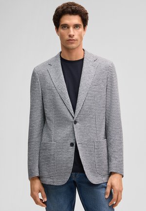 Mężczyzna w szarej marynarce w wzór pepitki, czarnej koszuli i niebieskich dżinsach, stoi na tle jednolitego jasnoszarego tła.