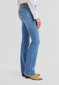 Asmuo dėvi šviesiai mėlynas Lee džinsus ir rudos spalvos smailius batus, stovintis prieš paprastą šviesiai pilką foną.