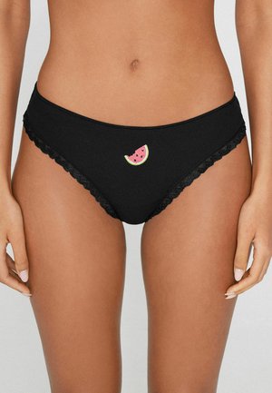 Sous-vêtement noir style bikini avec bordure en dentelle et une petite tranche de pastèque brodée au centre devant, porté par une personne debout.