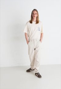 Tricou bej cu mâneci scurte și imprimeu grafic, asortat cu pantaloni evazați de culoare deschisă și adidași căprui cu dungi albe.