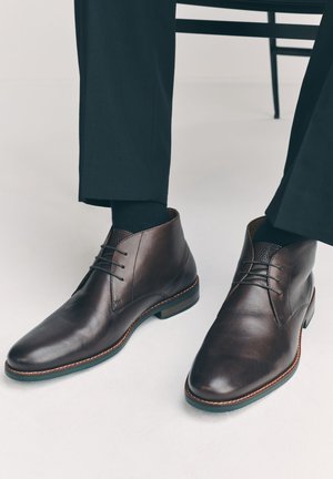Braune Leder-Schnürstiefeletten getragen mit schwarzen Socken und schwarzen Hosen, sitzend auf einem Stuhl mit sichtbaren Beinen.