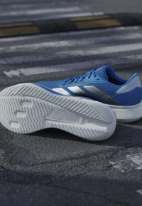 Chaussure de course Adidas bleu et blanc posée sur l’asphalte, semelle vers le haut, montrant une accroche texturée et les détails de la marque.