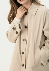 Beige trench coat, galléros nyakkivágással, hat nagy gombbal és egy zsebbel. Simább, textúrált anyagból készült és laza fazonú.