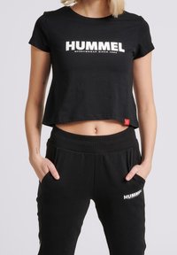 Černé bavlněné zkrácené tričko s krátkými rukávy a bílým textem "HUMMEL"; spárováno s černými tepláky se vzorem loga a elastickým pasem.