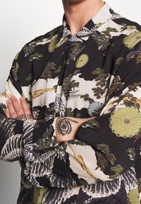 Koszula z długim rękawem, z czarnym tłem z zielonymi i kremowymi wzorami kwiatowymi oraz drzewnymi, z zapięciem na guziki z przodu i usztywnionym kołnierzykiem. Gładka tekstura.