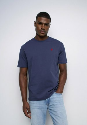 Homme portant un t-shirt bleu marine uni avec un petit logo rouge sur la poitrine et un jean bleu clair, debout contre un mur blanc.