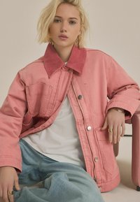 Lyserød denimjakke med fløjsfarvet krave, to forlommer og trykknapper. Modellen har lyseblå jeans og en hvid top på.