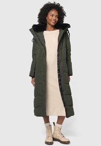 Olive green long puffer coat with a black faux fur hood, zip closure, side pockets, worn over a light beige dress and tan lace-up boots.