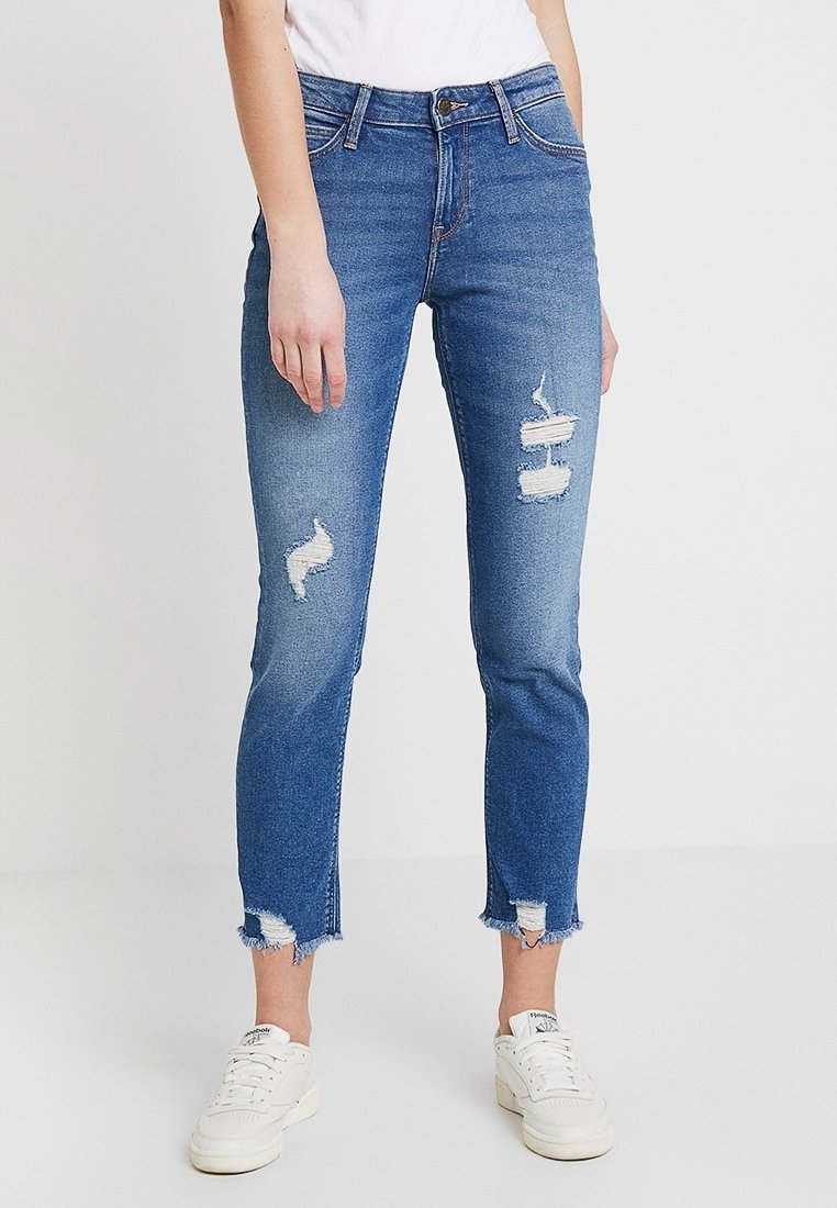 Femme portant un jean bleu déchiré et court avec des ourlets effilochés et des baskets blanches, se tenant contre un fond clair uni.