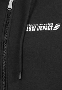 Black zip-up hoodie with a smooth texture, featuring white text "HIGH STANDARDS ALLOWED" and "LOW IMPACT" on the front. Zipper detail visible.