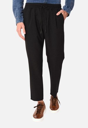 Joggers neri, tapered, con vita elastica e cordino; abbinati a sneakers marroni con suole e lacci bianchi.