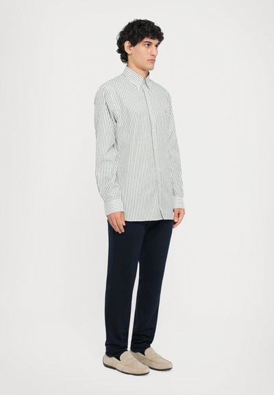 Cămașă button-up stripedă albă și bleumarin, cu mâneci lungi și guler. Asortată cu pantaloni bleumarin și pantofi slip-on bej, textură simplă.