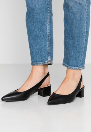 Osoba w niebieskich dżinsach i czarnych butach slingback na niskim, kwadratowym obcasie stoi na białej podłodze.