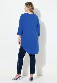 Tunic blu a manica lunga con orlo arrotondato, abbinata a jeans scuri con borchie laterali e sandali con tacco alto blu. Tessuto morbido.