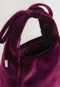 Geantă mică din catifea, de un violet intens, cu mâner noduros și capace metalice argintii la capete, deschisă cu o închizătoare cu nasture vizibilă în interior.