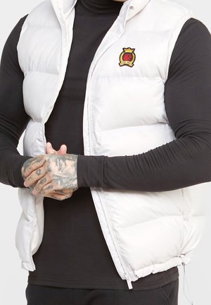 Mies, joka käyttää valkoista toppiliiviä mustan pitkähihaisen paidan päällä, kädet tatuoituina vyötäröllä ristissä, liivi puoliksi vetoketjulla suljettuna ja siinä vaakunamerkki.