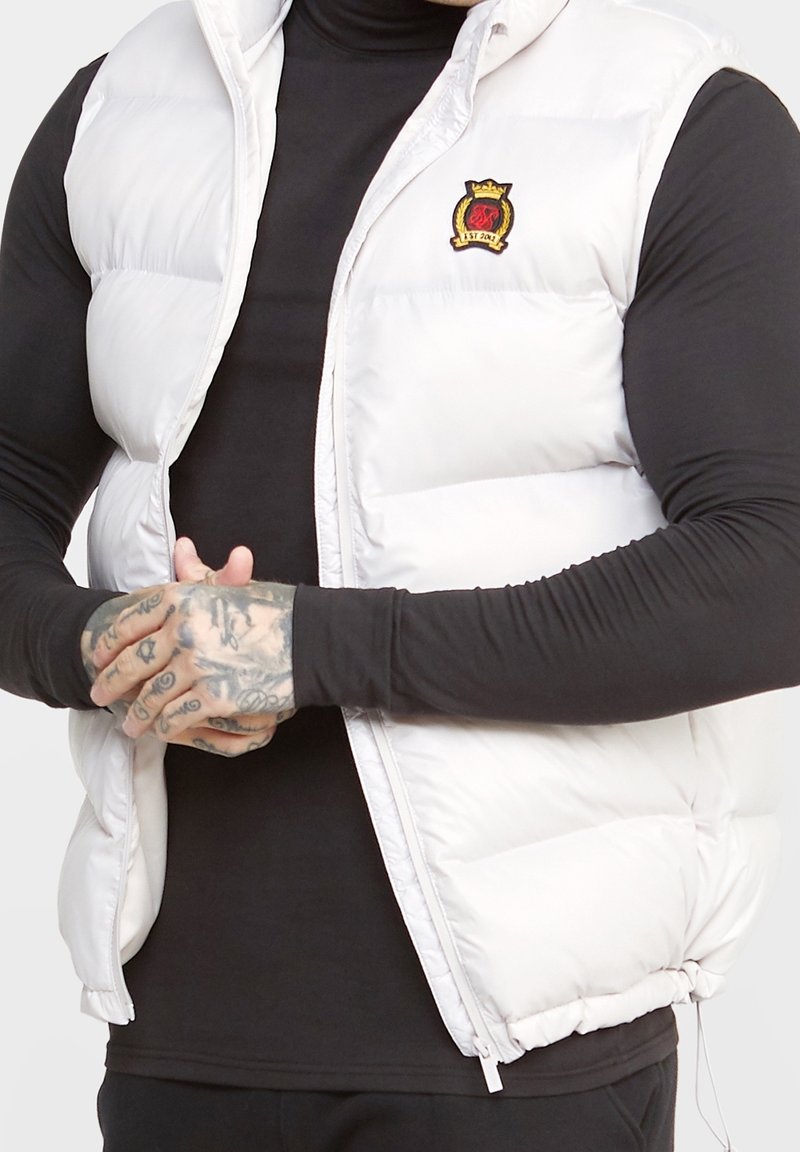 Mężczyzna ubrany w biały pikowany bezrękawnik na czarnej koszuli z długim rękawem, ręce z tatuażami splecione na wysokości talii, bezrękawnik zapięty do połowy, z logo herbu.