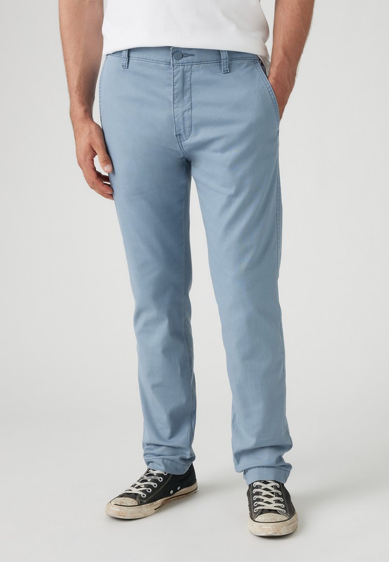 Világoskék, slim-fit pamut chinos, sima textúrával. Gombos záródással, övbújtatókkal és oldalsó zsebekkel rendelkezik. Fekete sportcipőkkel viselve.