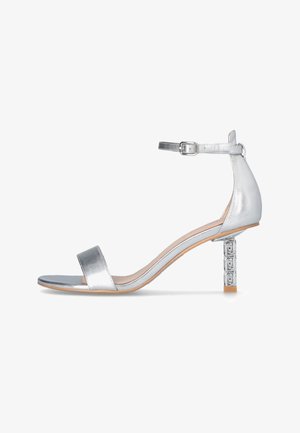 Sandale à talon argenté avec une finition métallique, une large bride avant, une bride à la cheville avec boucle et un talon bloc décoratif.