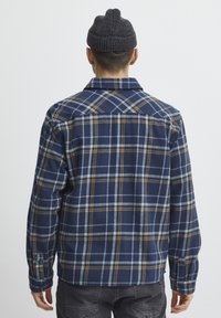 Camisa de franela de cuadro navy con patrones en azul, amarillo y blanco. Presenta un cuello, puños con botones y un corte relajado. Material cálido y texturizado.