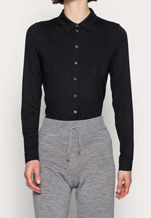 Vrouw draagt een aansluitend zwart overhemd met lange mouwen en knopen, ingestopt in hooggetailleerde grijze gebreide broek met een trekkoord in de taille.