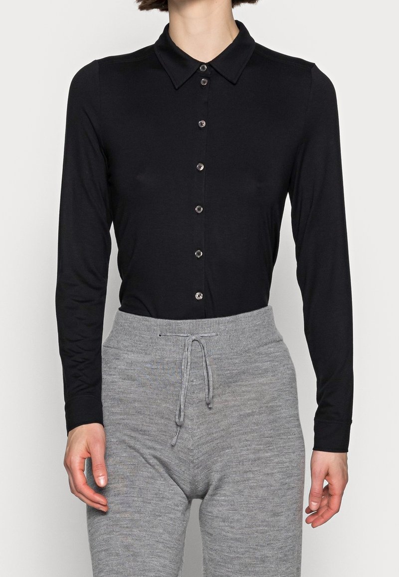 Femme portant une chemise noire ajustée à manches longues boutonnée, rentrée dans un pantalon en maille gris taille haute avec taille à cordon.