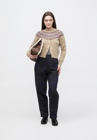 Beige ullkofta med rött mönster vid oket, vita knappar, mörka byxor med mönster i hög midja, håller en brun väska, svarta skor.