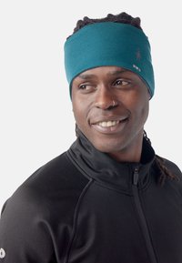 Bandeau teal en tissu doux, avec un petit logo et des accents colorés. Porté avec une veste noire zippée.