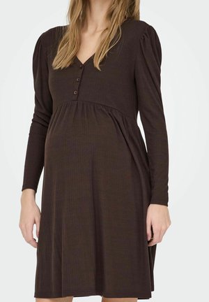 Kobieta w długim, ciemnobrązowym, prążkowanym sukienka z guzikami przy dekolcie w kształcie litery V i marszczonym detalem w talii.