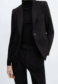 Person, der ein schwarzes maßgeschneidertes Blazer, einen schwarzen Rollkragenpullover und schwarze Hosen vor einem schlichten hellen Hintergrund trägt.