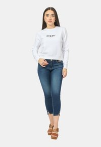 Felpa cropped bianca con logo "GUESS?", abbinata a jeans skinny blu scuro con dettagli in zip sul fondo e sandali con piattaforma color beige.