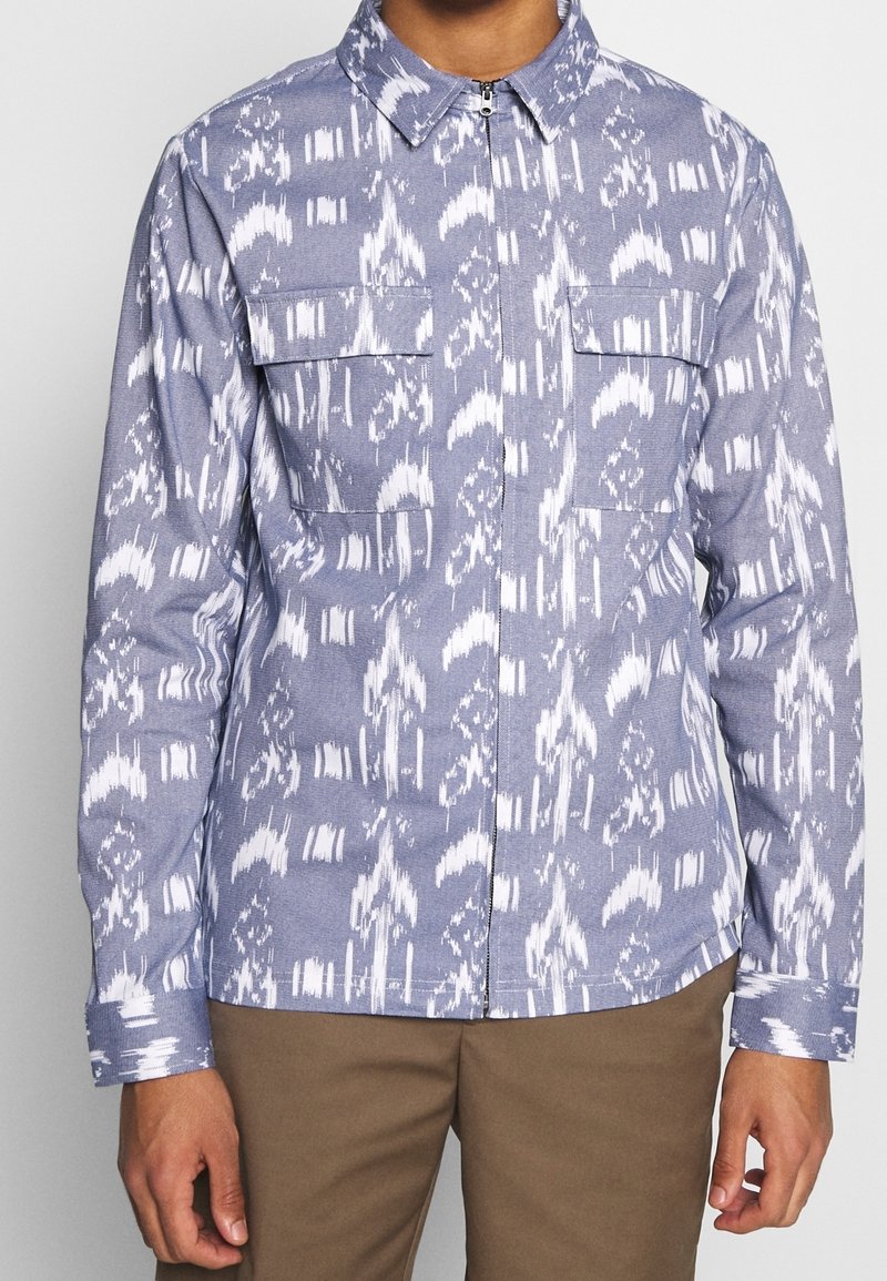 Man wearing blue and white patterned long-sleeve zip-up shirt with two chest pockets, paired with brown pants.