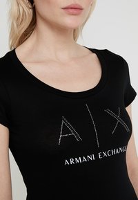 Černé tričko s krátkým rukávem a kulatým výstřihem, s stříbrně zdobeným logem "A|X" a textem "ARMANI EXCHANGE" na přední straně.