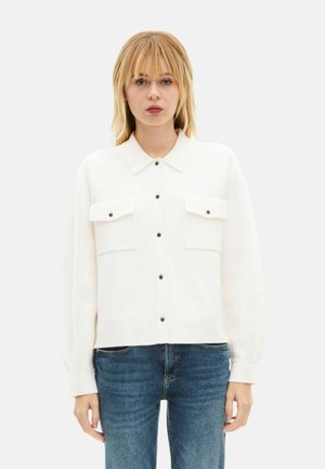 Camisa blanca abotonada con cuello, dos bolsillos delanteros y botones oscuros. Mangas largas con un ajuste ligeramente suelto. Combínala con vaqueros azules.