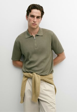 Junger Mann mit kurzen braunen Haaren, der ein grünes geripptes Poloshirt und beige Hosen trägt, mit einem beigen Pullover um die Taille gebunden.