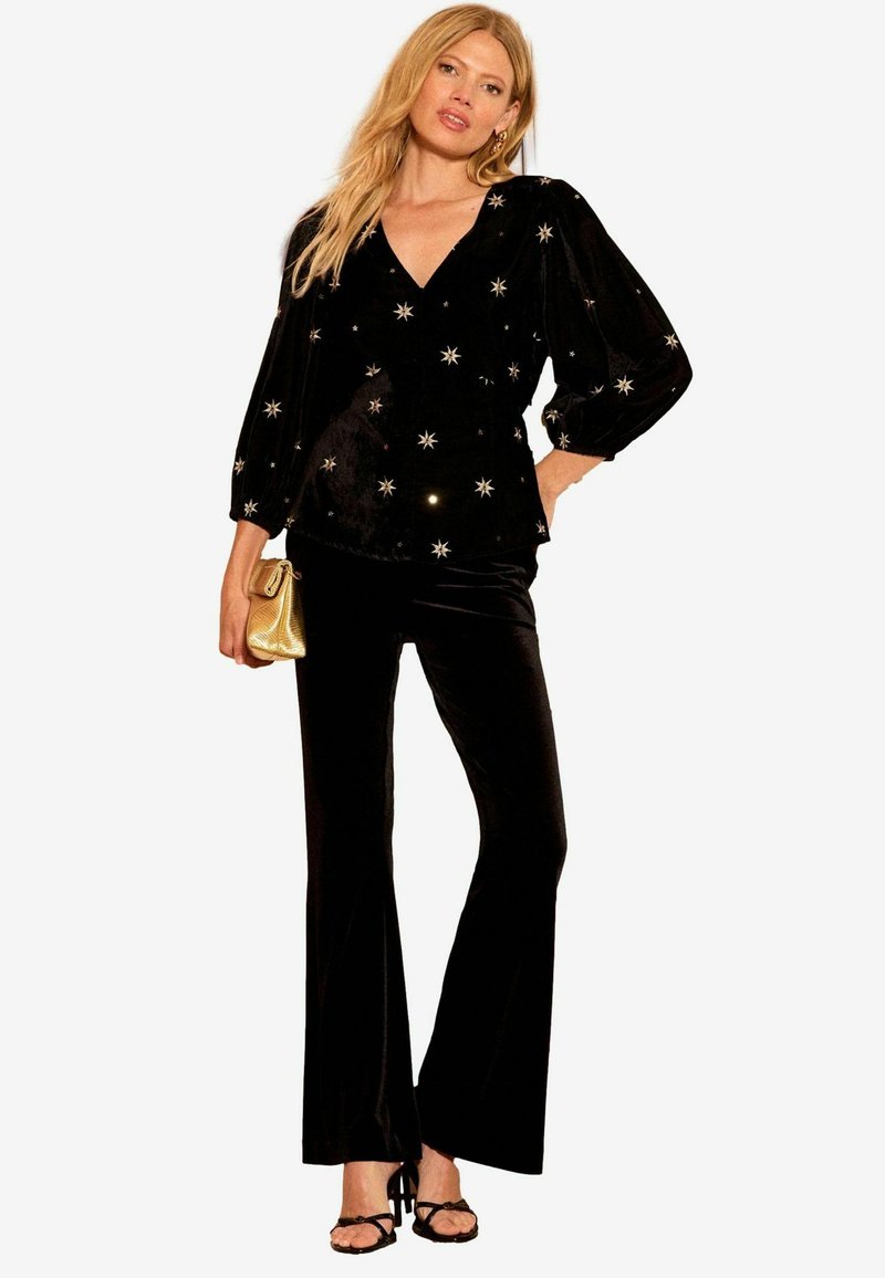 Top en velours noir avec un décolleté en V profond et des manches bouffantes, orné de embellissements en forme d'étoiles dorées. Associé à un pantalon noir évasé et une pochette dorée.