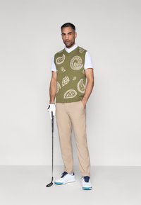 Žalios spalvos megztinis be rankovių su kreminiais paisley raštais, dėvimas ant balto marškinio. Aprangą papildo smėlio spalvos kelnės ir balti sportbačiai.